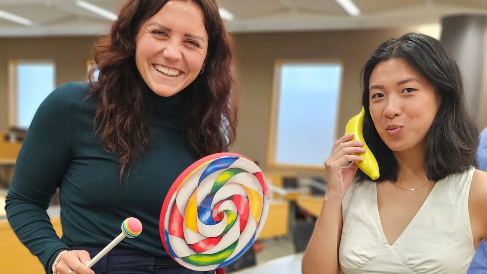 two students posing, one holding a tamporine shaped like a lollipop, the other holding a banana like it's a telephone